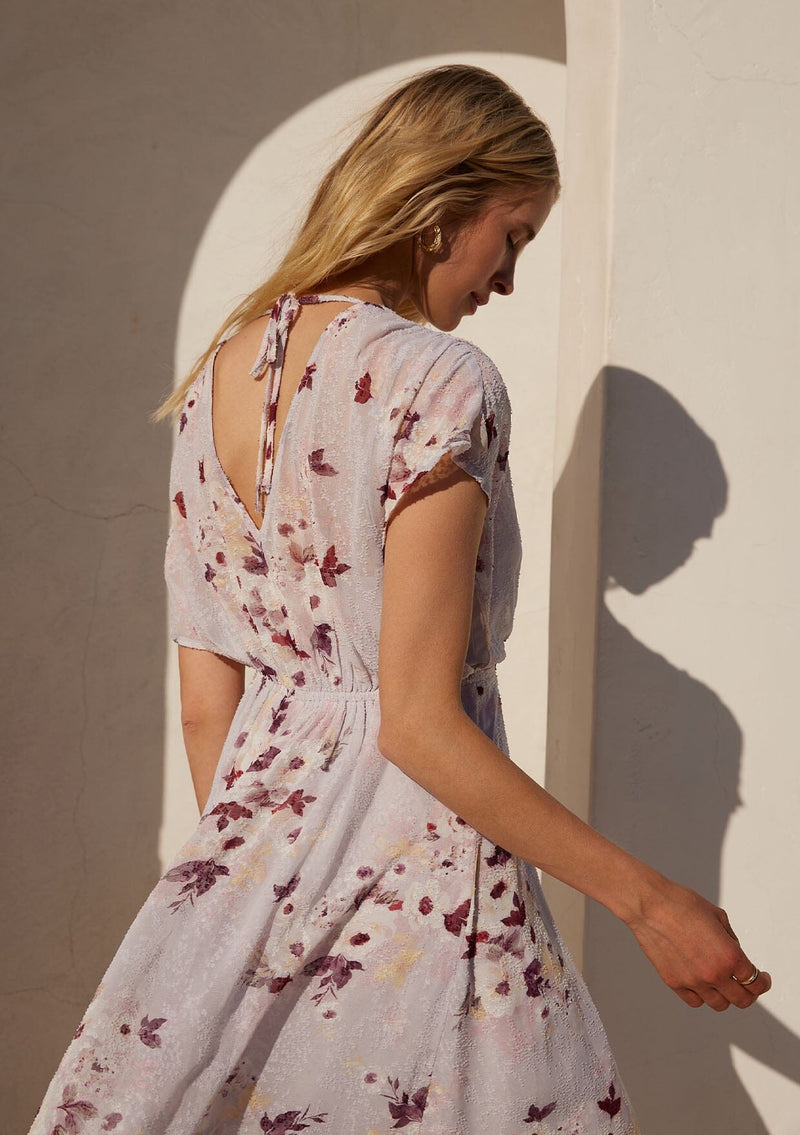 [Color: Dusty Blush/Wine] A close up back facing image of a blonde model standing outside wearing a bohemian maxi dress designed in a pink floral print and crafted from textured chiffon. With short dolman sleeves, a surplice v neckline, an open back with tie closure, and a flowy asymmetric hemline skirt.