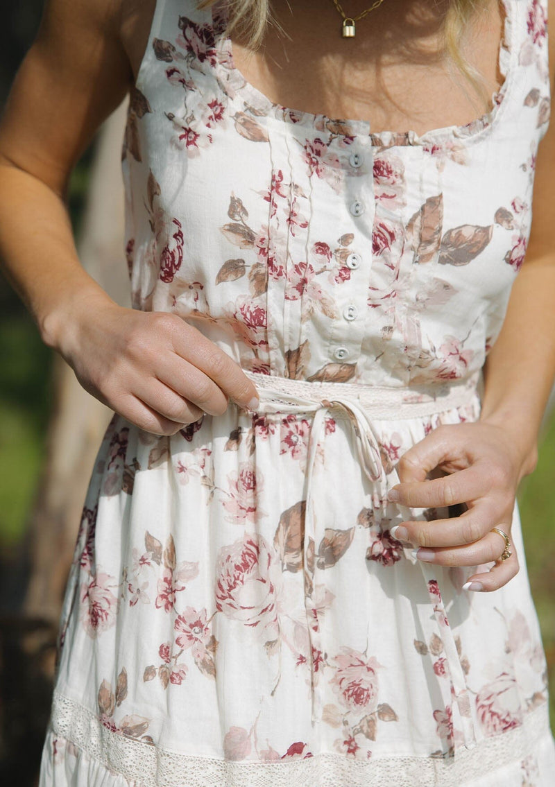 [Color: Natural/Dusty Wine] A front facing image of a brunette model wearing a sleeveless bohemian mid length dress in an off white and pink floral print. With a ruffle trimmed scoop neckline, a self covered button front top, a tiered skirt, lace trim, pintuck details, and a drawstring waist with ties.