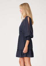 [Color: Ink Blue] A side facing image of a blonde model wearing a navy blue bohemian mini dress in embroidered chiffon. With long split sleeves, a ruffle trimmed tiered skirt, a smocked elastic waist, a v neckline, and an open back with tassel tie closure. Perfect for weddings or date nights.