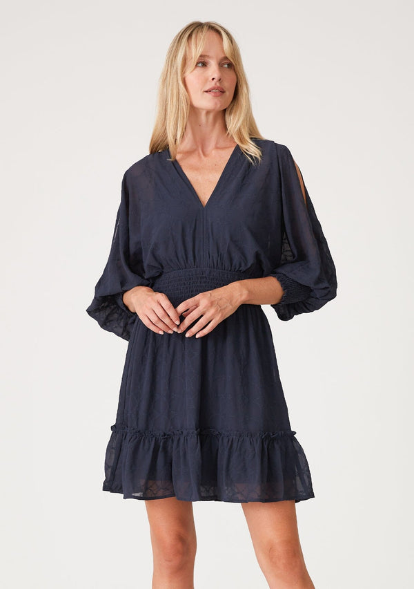[Color: Ink Blue] A front facing image of a blonde model wearing a navy blue bohemian mini dress in embroidered chiffon. With long split sleeves, a ruffle trimmed tiered skirt, a smocked elastic waist, a v neckline, and an open back with tassel tie closure. Perfect for weddings or date nights.