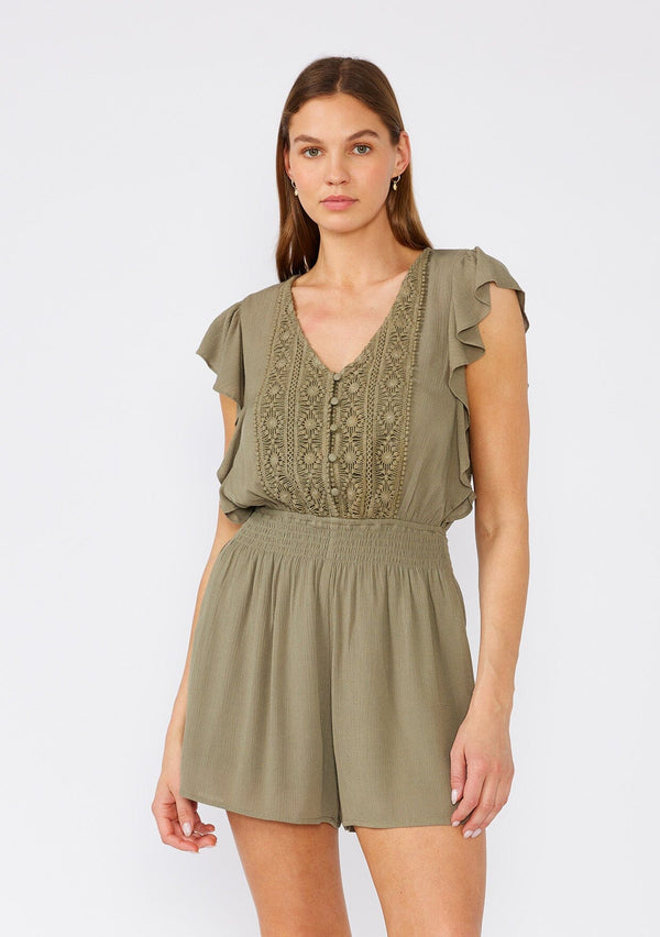 [Color: Olive] A front facing image of a brunette model wearing an olive green bohemian short romper. With short flutter sleeves, a v neckline, a crochet top, a self covered button front top, and a smocked elastic waist. 