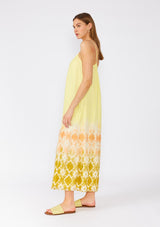 [Color: Lemon/Coral] A side facing image of a brunette model wearing a bohemian summer cotton maxi dress in a yellow tie dye print. With adjustable spaghetti straps, a scooped neckline with ruffled trim, a roomy fit, and side pockets. 