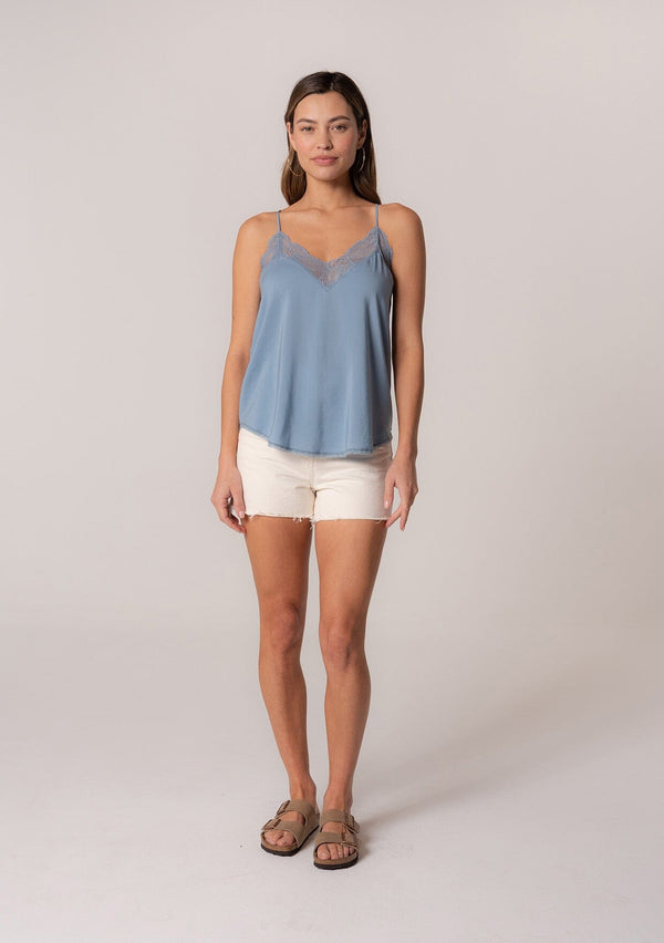 [Color: Denim] A front facing image of a brunette model wearing a blue camisole tank top. With spaghetti straps, a v neckline, lace trim, a racerback, and a relaxed fit. 