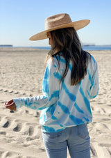 [Color: White Lagoon] A woman on the beach wearing a soft and cozy tie dye sweatshirt. This cozy pullover features long raglan sleeves and a classic crew neckline. A great option for working from home.