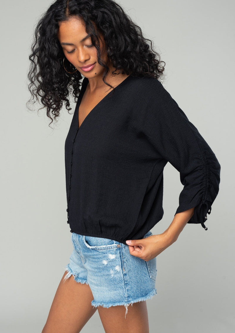 [Color: Black] A side facing image of a brunette model wearing a classic black bohemian top with a self covered button front, a v neckline, and three quarter length sleeves with a gathered sleeve detail and adjustable ties. 