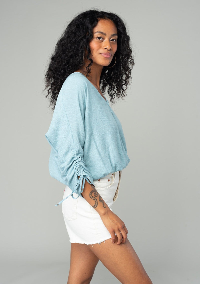 [Color: Dusty Blue] A side facing image of a brunette model wearing a classic dusty blue bohemian top with a self covered button front, a v neckline, and three quarter length sleeves with a gathered sleeve detail and adjustable ties. 