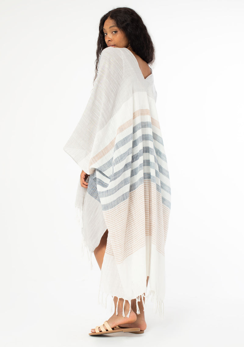 [Color: Neutral Combo] A side facing image of a black model with long wavy hair wearing a cotton bohemian kimono with neutral blue and white white detail and fringe hemline. Model is looking over her shoulder. 