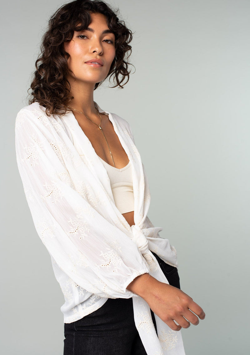 [Color: Ivory] A side facing image of a brunette model wearing an ivory white bohemian embroidered chiffon kimono top. With long sleeves and a tie front that can be styled in multiple ways. 