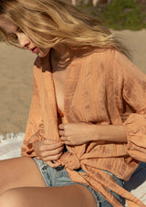 [Color: Spice] A close up side facing image of a blonde model at the beach wearing a best selling bohemian tie front resort top in a light orange shadow stripe. With voluminous long sleeves and a tie front detail that can be styled in multiple ways.