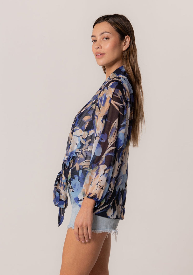 [Color: Navy/Light Blue] A side facing image of a brunette model wearing a sheer chiffon tie front top in a blue floral print. With long sleeves, a deep v neckline, a flowy relaxed fit, and a tie front waist detail. 