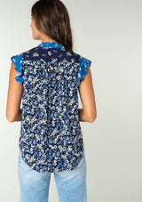 [Color: Navy/Coral] A back facing image of a brunette model wearing a navy blue and coral mixed floral print bohemian top. With a button front, short flutter sleeves, and neck ties. 