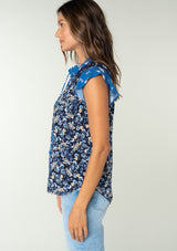 [Color: Navy/Coral] A side facing image of a brunette model wearing a navy blue and coral mixed floral print bohemian top. With a button front, short flutter sleeves, and neck ties. 