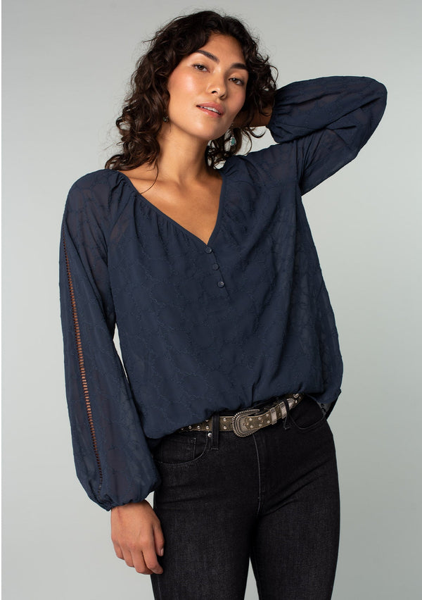 [Color: Ink Blue] A front facing image of a brunette model wearing a navy blue bohemian peasant top in embroidered chiffon. With long sleeves, lattice trim, a v neckline, a button front, and a flowy relaxed fit. 
