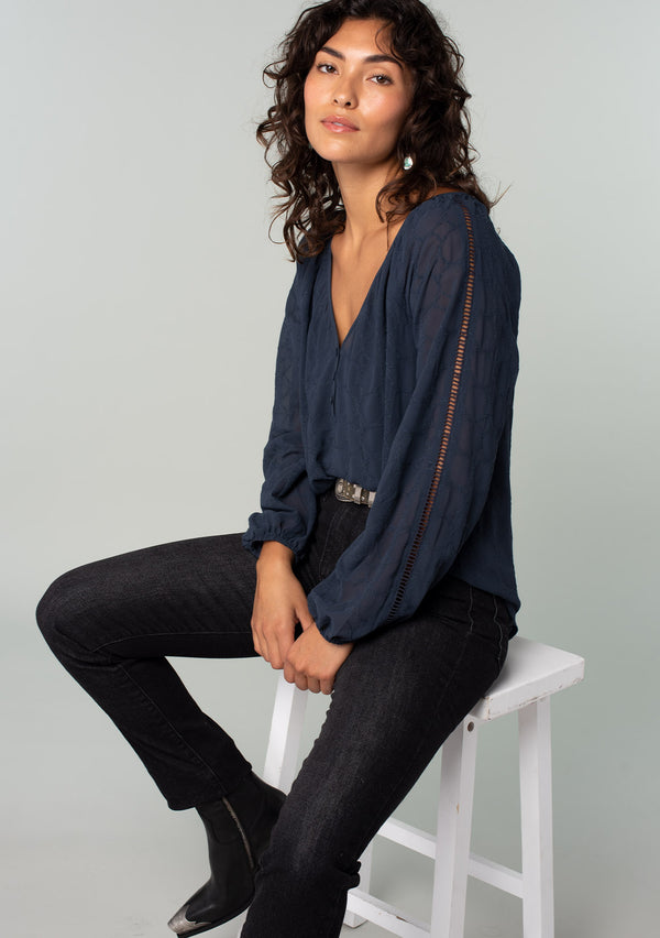 [Color: Ink Blue] A sitting side facing image of a brunette model wearing a navy blue bohemian peasant top in embroidered chiffon. With long sleeves, lattice trim, a v neckline, a button front, and a flowy relaxed fit. 
