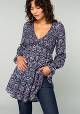 [Color: Navy/Tan] A front facing image of a brunette model wearing a navy blue and tan paisley print tunic top. A vintage inspired top with long sleeves, a smocked bodice, an empire waist, and a deep v neckline.