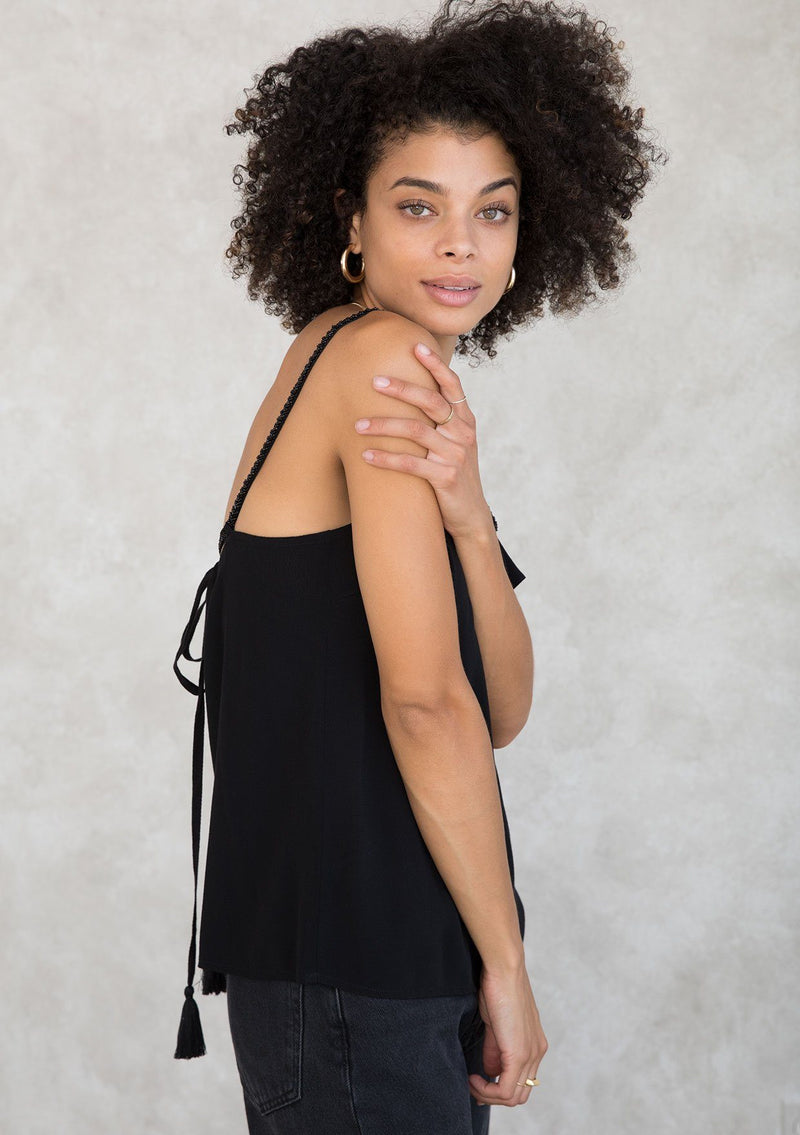 [Color: Black] A model wearing a Holiday ready black camisole. For your next special occasion, featuring a ruffle trimmed front and a beaded accent neckline with long beaded straps that tie in the back. 