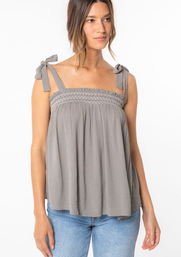 [Color: Cement] A woman wearing a grey crinkle gauze flowy bohemian tie shoulder tank top with smocked neckline. 