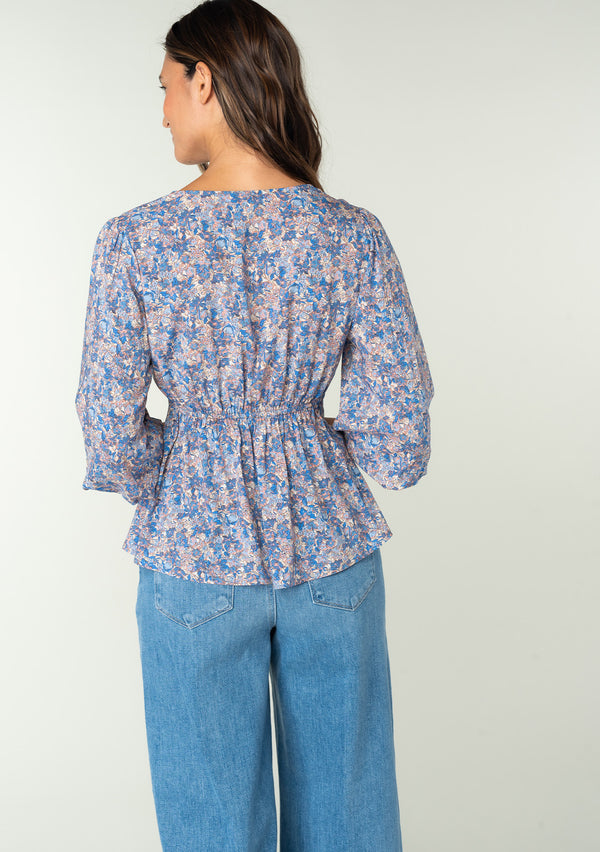 [Color: Blue/Coral] A back facing image of a brunette model wearing a classic bohemian peplum blouse in a blue and coral floral print. With long three quarter length sleeves, a v neckline, and a button front. 