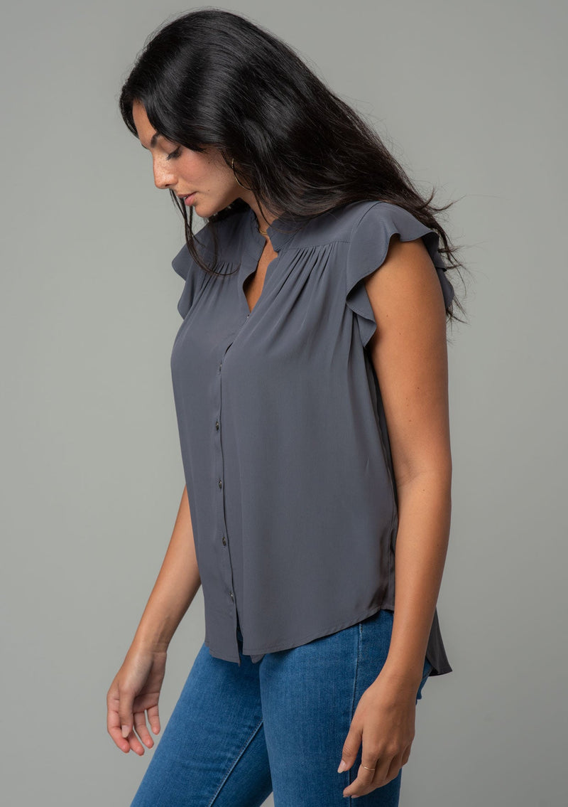 [Color: Pewter] A side facing image of a brunette model with long dark wavy hair wearing a classic bohemian short flutter sleeve button front top in a dark grey silky crepe. 