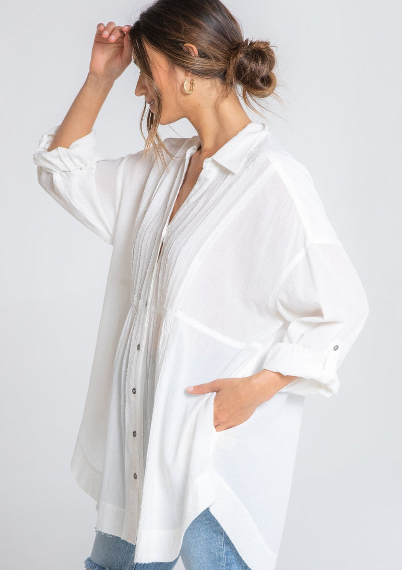 [Color: Off White] A model wearing a relaxed cotton tunic shirt with a button front, long rolled sleeves, and pleated details. 