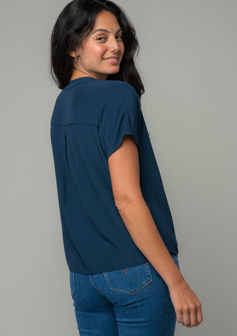 [Color: Midnight] A side facing image of a brunette model wearing a soft and silky navy blue short sleeve top with a button front and tie waist detail. 