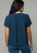 [Color: Midnight] A back facing image of a brunette model wearing a soft and silky navy blue short sleeve top with a button front and tie waist detail. 