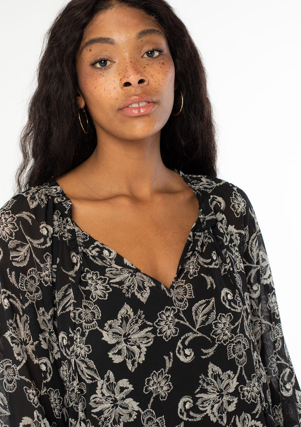 [Color: Black/Natural] A close up front facing image of a black model wearing a sheer chiffon bohemian blouse in a black and natural floral print. With long sleeves, a flowy silhouette, and a split v neckline with tassel neck ties. 