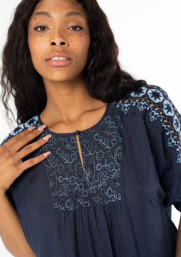 [Color: Navy] A close up front facing image of a black model wearing a navy blue cotton bohemian top with half length dolman sleeves and a contrast light blue embroidered detail along the sleeve and front bib. 
