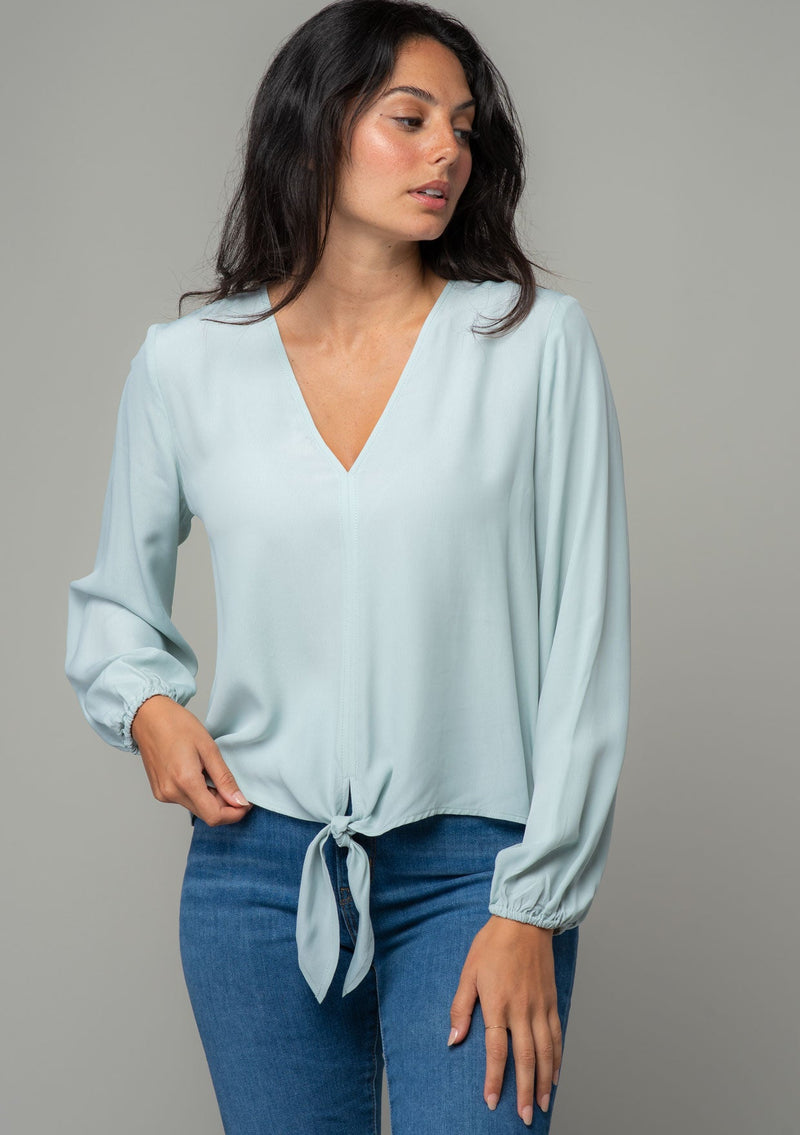 [Color: Sky] A front facing image of a brunette model wearing a soft and silky light blue crepe long sleeve top with a tie front detail. 