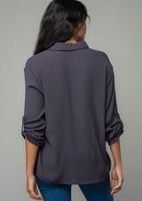 [Color: Charcoal] A half body back image of a brunette model wearing a relaxed fit charcoal grey button front shirt in a lightweight crepe. With long rolled sleeves, a button tab sleeve closure, a front placket, and a collared neckline. 