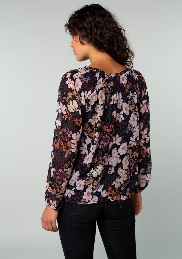 [Color: Black/Dusty Rose] A back facing image of a brunette model wearing a chiffon bohemian blouse in a black and purple floral print. With long sleeves and a smocked elastic round neckline.