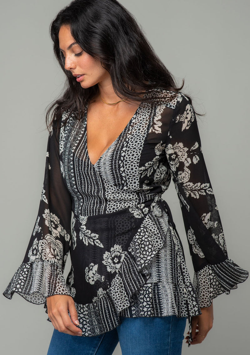 [Color: Black/Cream] A half body front facing image of a brunette model wearing a bohemian hippie wrap top in a mixed black and cream floral print. With long bell sleeves and a ruffled wrist cuff, a deep v neckline, and a side tie closure. 