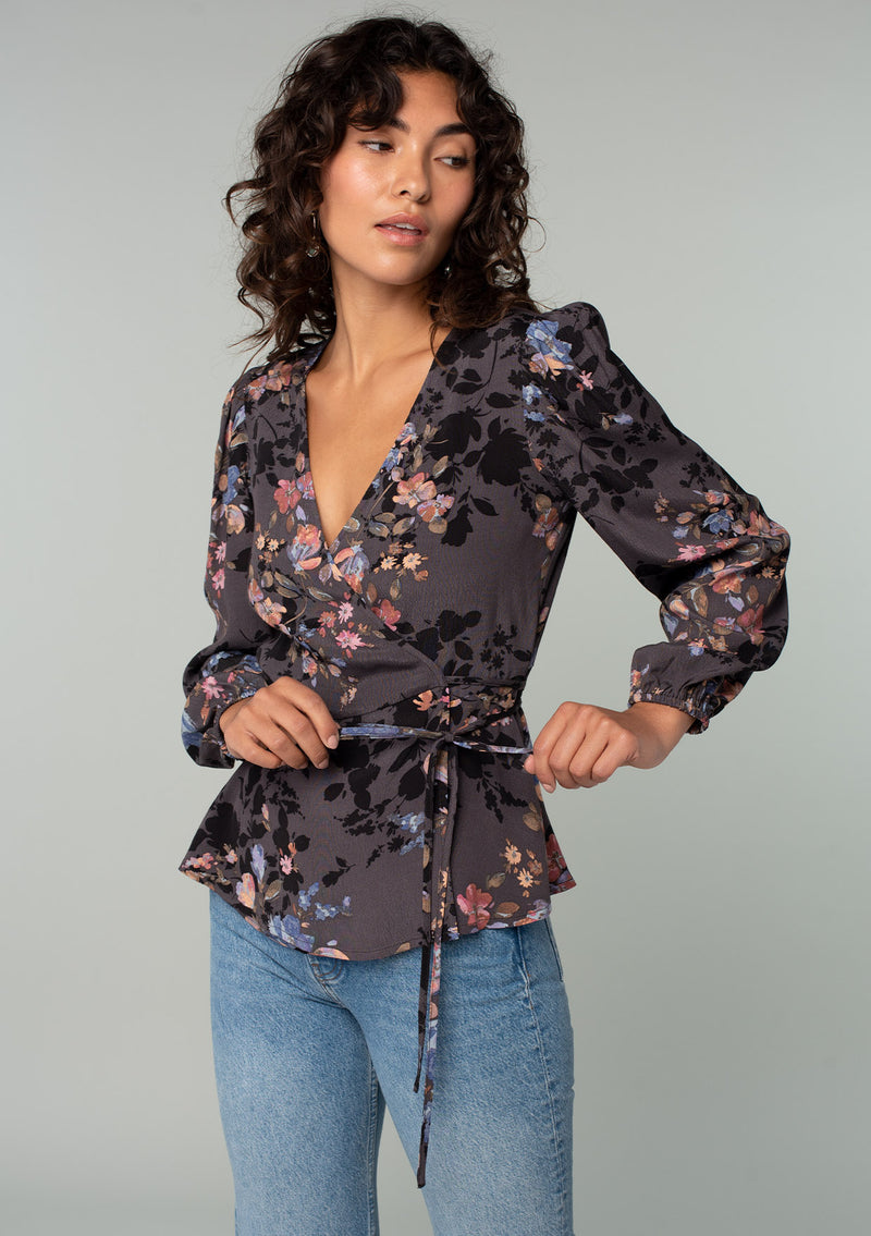 [Color: Grey/Dusty Blue] A half body front facing image of a model wearing a bohemian wrap top in a dark grey and dusty blue floral print. With long sleeves and a side tie closure. 