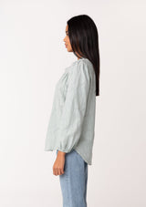 [Color: Sage] A side facing image of a brunette model wearing a classic bohemian cotton sage green button front shirt in a textured jacquard. With long sleeves and a ruffled neckline.