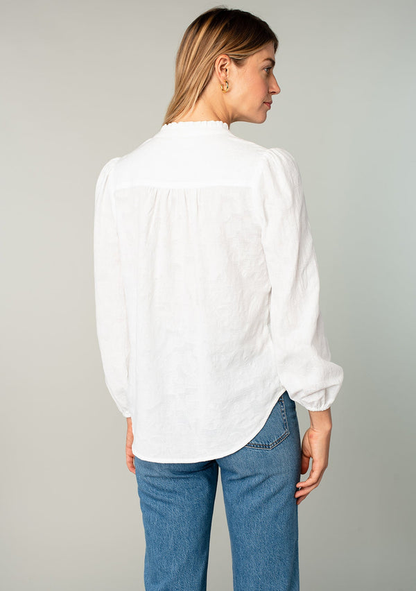 [Color: White] A back facing image of a blonde model wearing a classic bohemian cotton white button front shirt in a textured jacquard. With long sleeves and a ruffled neckline. 