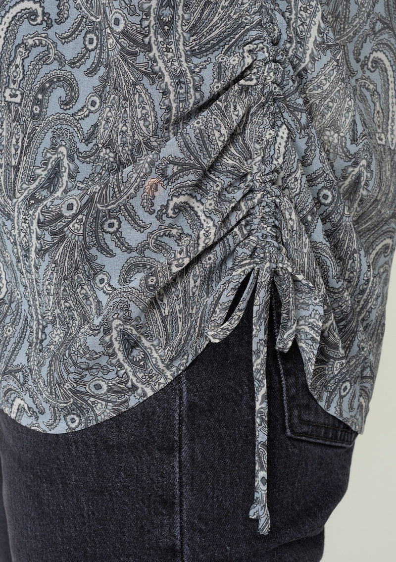 [Color: Grey/Natural] A close up side facing image of a brunette model wearing a sheer chiffon button front shirt in a grey and natural paisley print. With a collared neckline, long sleeves with a button wrist cuff, and an adjustable side gathered waist detail with ties. 
