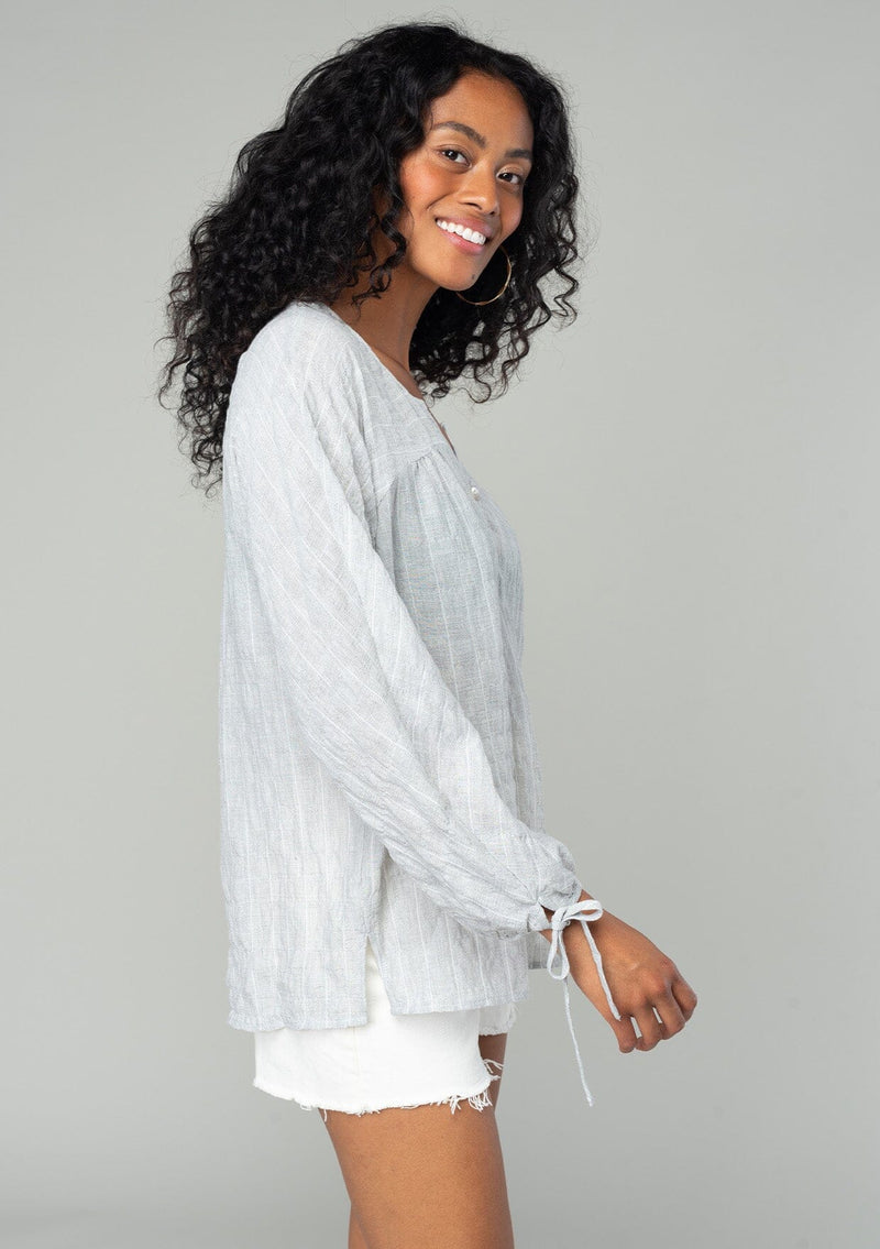 [Color: Heather Grey] A side facing image of a brunette model wearing a sheer cotton bohemian top in a heather grey striped jacquard. With long sleeves, a button front, and tie wrist cuffs.