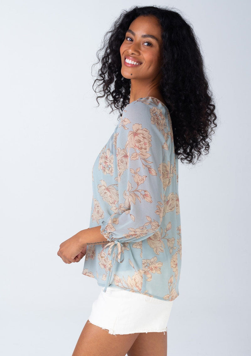 [Color: Dusty Blue/Natural] A side facing image of a brunette model wearing a sheer chiffon spring bohemian blouse in a dusty blue and natural floral print. With three quarter length sleeves, adjustable tie cuffs, a v neckline, and a flowy relaxed fit. 