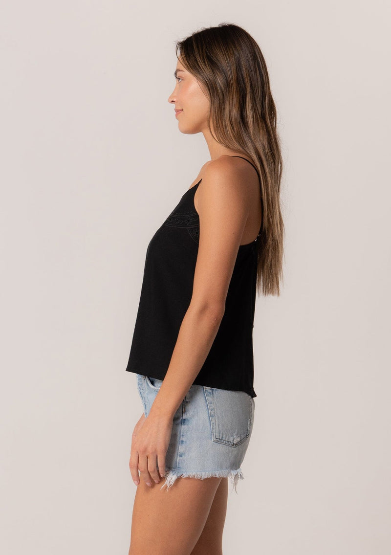 [Color: Black] A side facing image of a brunette model wearing a black bohemian spring camisole tank top with embroidered detail, adjustable spaghetti straps, a scooped neckline, and a self covered button up back detail.