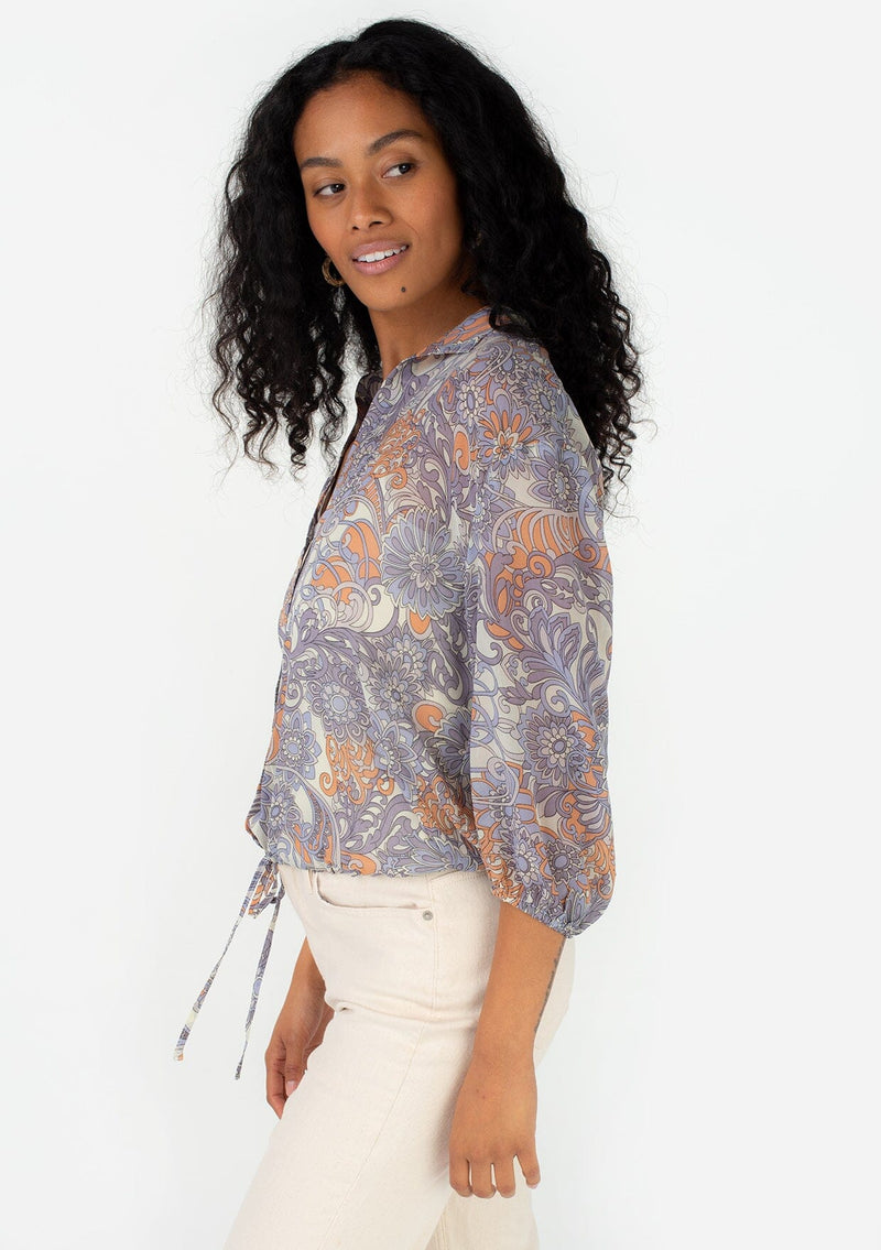[Color: Ivory/Dusty Plum] A side facing image of a brunette model wearing a sheer chiffon bohemian blouse in a retro inspired purple floral print. With three quarter length sleeves, a collared neckline, a button front, and a drawstring waist with adjustable tie. 