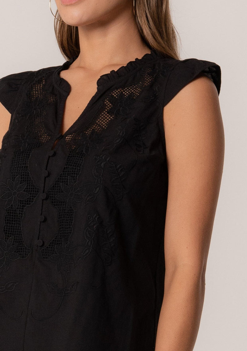 [Color: Black] A close up front facing image of a brunette model wearing a bohemian cotton spring top in a black colorway. With short cap sleeves, a self covered button front, a ruffled neckline, a v neckline, and embroidered detail.