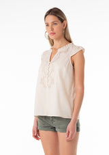 [Color: Natural] An angled front facing image of a blonde model wearing a bohemian cotton spring top in a natural colorway. With short cap sleeves, a self covered button front, a ruffled neckline, a v neckline, and embroidered detail. 