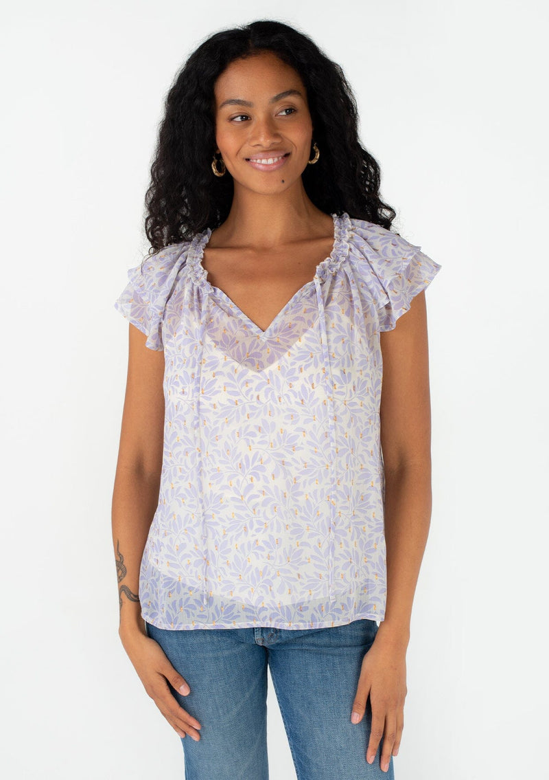[Color: Natural/Lilac] A front facing image of a brunette model wearing a pretty spring blouse in a metallic clip dot chiffon purple leaf print design. With short flutter sleeves, a ruffled neckline, a split v neckline with ties, and a relaxed fit. 