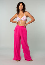 [Color: Fuchsia] A full body front facing image of a brunette model wearing a bright pink boho lounge pant in natural cotton gauze. With a wide leg, side pockets, and a smocked elastic waistband, these bohemian beach pants are prefect for summer days.