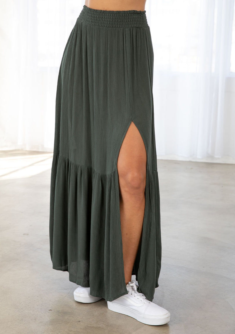 [Color: Military] A model wearing a military green maxi skirt. With a smocked elastic waist, a side slit, a tiered skirt, and a flowy silhouette. 