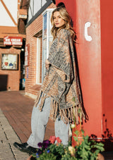 [Color: Coco/Charcoal] A blond woman outside wearing an ultra soft bohemian knit poncho sweater. Featuring a flirty fringed hemline, a blanket stitch detail, and an open front.