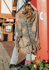 [Color: Coco/Charcoal] A blond woman outside wearing an ultra soft bohemian knit poncho sweater. Featuring a flirty fringed hemline, a blanket stitch detail, and an open front.