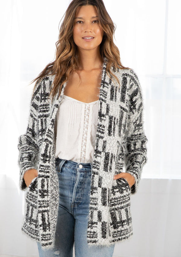 [Color: Silver/Black] A model wearing a fuzzy black and white abstract stripe cardigan sweater. A mid length style featuring long sleeves, side pockets, and an open front. 