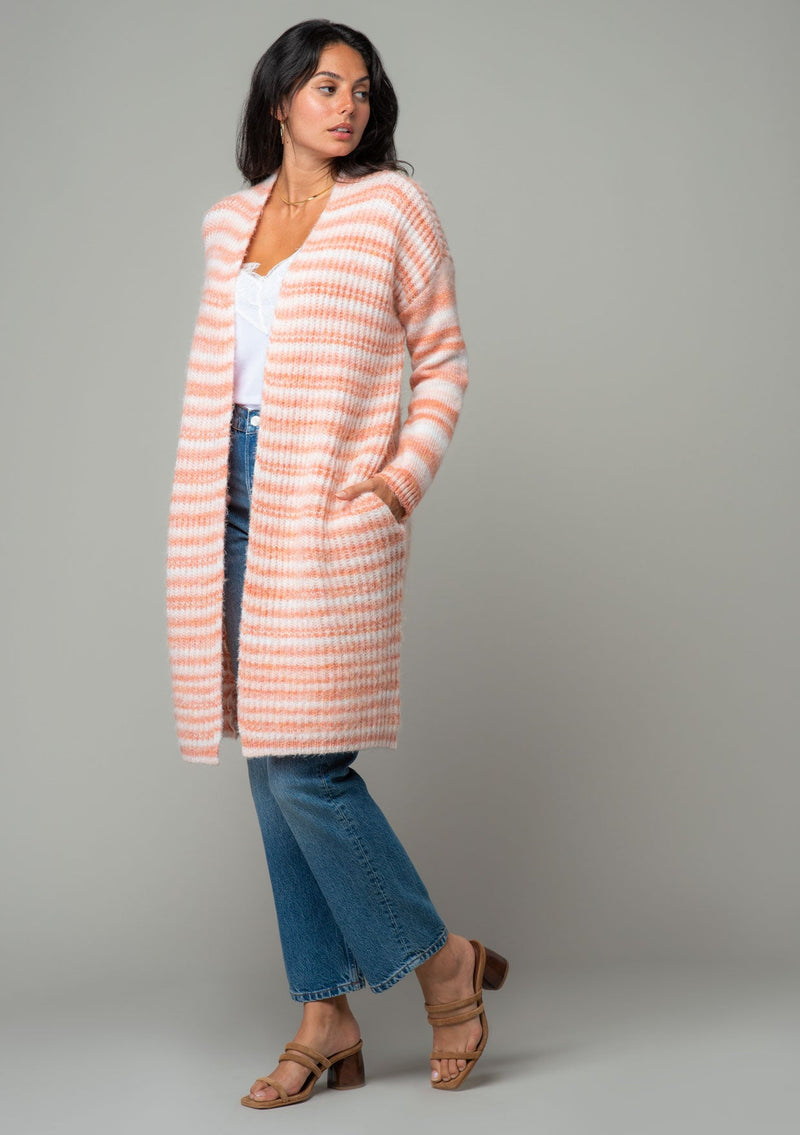 [Color: Ivory/Peach] A side facing image of a brunette model wearing a white and peach striped long cardigan. With an open front, contrast striped long sleeves, a shawl collar, and side pockets. 