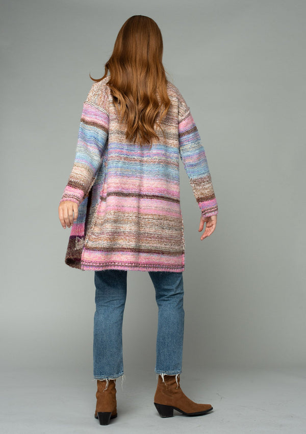 [Color: Pink Multi] A back facing image of a red headed model wearing a multi color pink patchwork knit cardigan. A mid length bohemian cardigan with long sleeves, an open front, and exposed seam details. 